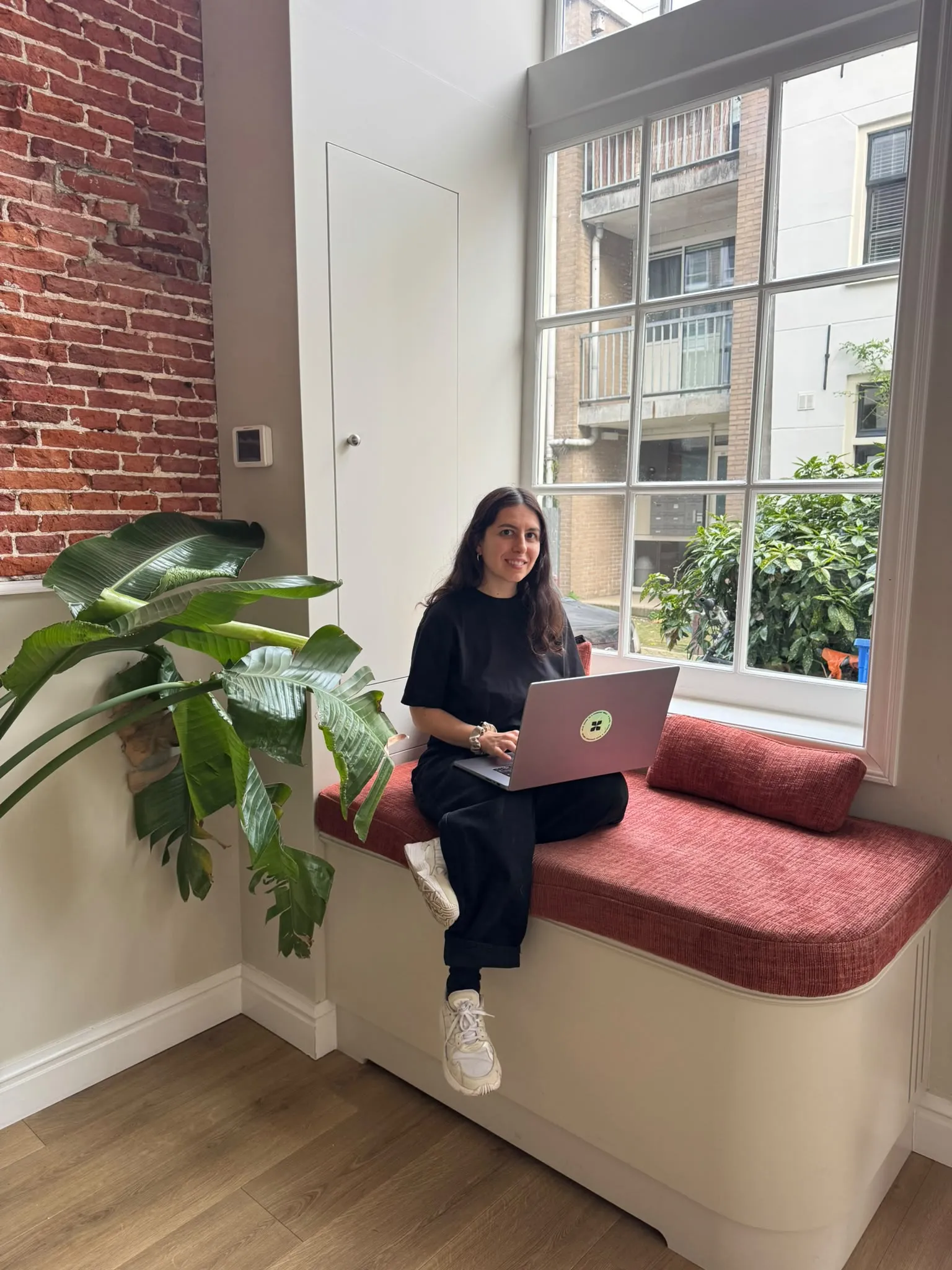 Antonella Pitti sitting with her laptop beside a sunny window and houseplants.